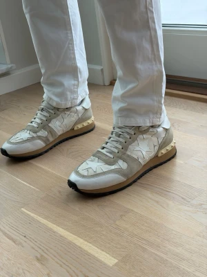 Valentino Rockrunner beige sneakers - Snygga Valentino Rockrunner sneakers i beige och vitt med ikoniska stjärndetaljer på sidan. Skorna har snörning, platt sula och guldfärgade nitar på hälen. Materialmix av mocka, skinn och textil för en lyxig känsla.