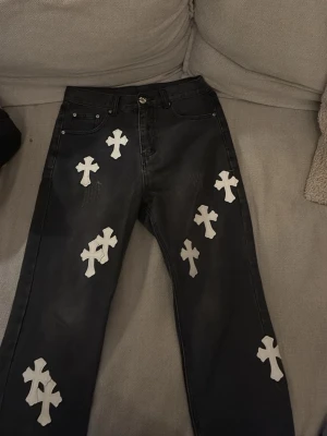 Svarta jeans med vita korsdetaljer - Unika svarta jeans med flera vita korsapplikationer framtill och baktill. Rak passform och klassisk femficksdesign. Jeansen har en edgy vibe och sticker verkligen ut i mängden. Perfekta för dig som vill ha något annorlunda i garderoben.