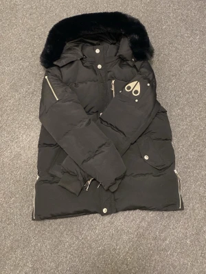 Svart dunjacka Moose Knuckles päls - Svart dunjacka från Moose Knuckles med pufferstil, päls på huvan och silverfärgade dragkedjor. Jackan har logga på ärmen, flera fickor med knappar och dragkedja framtill. Perfekt för vintern och riktigt varm.