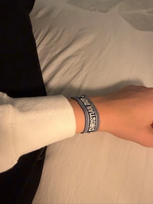 Dior armband - Snyggt blått tygarmband från Christian Dior med texten 'J'Adior' broderad i vitt. Armbandet har justerbar knytning med tofsar i ändarna och en cool, avslappnad stil. Perfekt accessoar för att lyfta din outfit med en exklusiv touch.