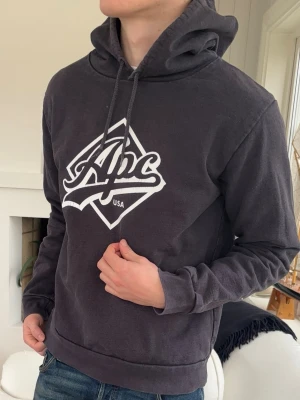 Mörkgrå hoodie från Apc  - Snygg mörkgrå hoodie från Apc USA med stor vit logga på bröstet. Klassisk modell med huva och dragsnören, samt ribbade muddar vid ärmar och nederkant. Perfekt för en avslappnad och trendig stil. Tillverkad i mjukt bomullsmaterial.