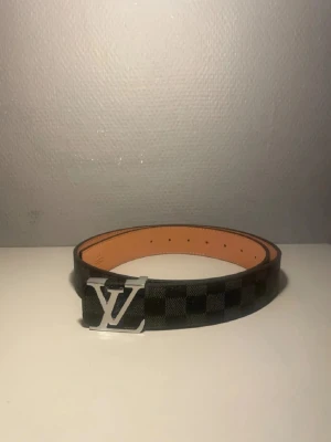 Iouis Vuitton rutigt bälte - Snyggt bälte från Iouis Vuitton med klassiskt brunt och svart rutmönster och stor IV-logga i silver på spännet. Bältet har en stilren design och insidan är ljusbrun. Perfekt accessoar för att lyfta din outfit.