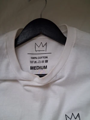 Vit Hov1 tour t-shirt - Säljer en vit t-shirt från Hov1 med tryck från deras 'Vi Dansar Vidare I Livet'-turné på ryggen och en liten krona broderad fram. T-shirten är i 100% bomull och har klassisk passform med korta ärmar. Perfekt för dig som gillar svensk musik och vill ha en unik merch!