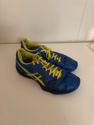 Blåa Asics inomhusskor med gula detaljer - Säljer ett par sportiga Asics inomhusskor i blått med gula snören och detaljer. Skorna har låg profil, slitstark sula och ventilerande meshmaterial. Perfekta för snabba rörelser på planen och ger bra grepp. Snygg design med Asics-logga på sidan och sulan. Slitna vid hälen, därav billigt pris💙