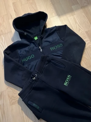 Svart set från Hugo Boss med huva - Snyggt svart set från Hugo Boss med både hoodie och byxor. Hoodien har dragkedja, huva och gröna logotexter på bröstet. Byxorna har resår i midjan, snörning och grönt Boss-tryck på benet. Perfekt för en avslappnad och trendig look.