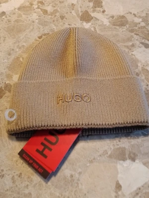 Beige ribbstickad mössa från HUGO Boss - Fin Hugo mössa som original märket är Hugo Boss. Den är helt oanvänd med alla etiketter på. Alla qr koder elder till deras hemsida så att den är riktig. Mössa är fin och stretchig om tillåter dig att ha en stor komfort. Skriv till mig om det är frågor!