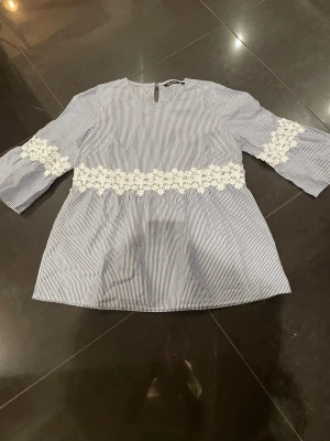 Blårandig blus med spetsdetaljer - Snygg blus med tunna blå och vita ränder samt vita spetsdetaljer i midjan och på ärmarna. Blusen har trekvartsärm och en avslappnad passform. Perfekt till jeans eller kjol för en trendig look.