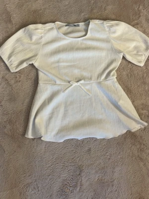 Vit topp med knyt och kort ärm - Snygg vit topp med rund halsringning, korta puffärmar och ett smalt knytband i midjan som ger en feminin siluett. Materialet har en lätt ribbad struktur och toppen har en något utsvängd nederdel. Perfekt att styla med jeans eller kjol.