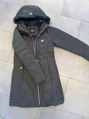 Mörkgrå lång dunjacka från Áhkká - Lång, mörkgrå dunjacka från Áhkká med huva och dragkedja framtill. Jackan har quiltad design, två fickor med dragkedja och diskret logga på bröstet. Perfekt för kalla dagar och har en snygg, figurnära passform.