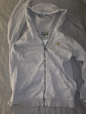 Grå zip hoodie från Lyle & Scott - Snygg grå hoodie med dragkedja från Lyle & Scott. Klassisk design med huva, fickor framtill och den ikoniska gula loggan på bröstet. Perfekt för en avslappnad stil och enkel att matcha med andra plagg. Tillverkad i mjukt bomullsmaterial.