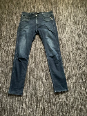 Mörkblå Replay Anbass slim jeans - Snygga mörkblå jeans från Replay, modell Anbass med slim fit. Klassisk femficksdesign och diskreta slitningar framtill. Jeansen har dragkedja och knapp i midjan, samt detaljerad söm vid fickorna. Perfekta för en trendig och stilren look. Var inte rädd för att skicka om du har några frågor eller vill ha fler bilder!