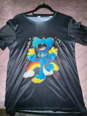 Svart t-shirt med färgglatt tryck - Cool svart t-shirt med ett stort färgglatt tryck av en tecknad blå figur, regnbåge och stjärnor på framsidan. T-shirten har rund hals och korta ärmar. Perfekt för dig som gillar lekfulla och unika motiv.