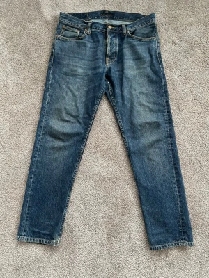 Blå jeans från Nudie Jeans - Säljer ett par klassiska blå jeans från Nudie Jeans med raka ben och fem fickor. Jeansen har en snygg tvättad look och kontrastsömmar samt Nudie-logga på bakfickan. Jeansen har riktigt go fade.Vid frågor är det bara att slänga iväg ett meddelande!