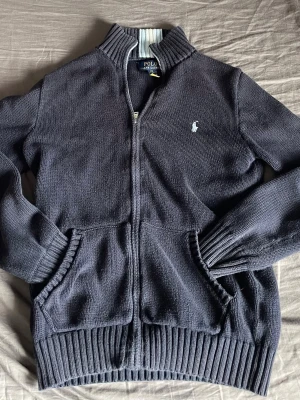 Mörkblå stickad ziptröja Polo Ralph Lauren - Mörkblå stickad ziptröja från Polo Ralph Lauren med ribbad krage, muddar och nederkant. Tröjan har två fickor framtill och den klassiska broderade loggan på bröstet. Perfekt för lager på lager och chill stil.