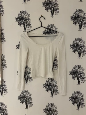 Vit ribbad långärmad topp - Snygg vit ribbad topp med lång ärm och lätt croppad passform. Toppen har en bred, rundad urringning med subtil spetskant som ger en feminin touch. Perfekt att styla med jeans eller kjol för en trendig look.