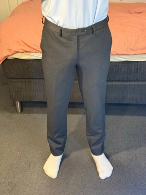 Mörkgrå kostymbyxor med rak passform - Snygga mörkgrå kostymbyxor med klassisk rak passform och bälteshällor. Byxorna har två bakfickor med knapp och stängs med knapp och dragkedja fram. Perfekta till skjorta eller kavaj för en clean look.