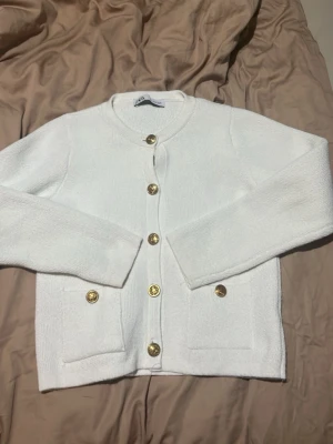 Vit kofta med guldfärgade knappar från Zara - Säljer en stilren vit kofta från Zara med rund halsringning och långa ärmar. Koftan har snygga guldfärgade knappar framtill och två fickor. Perfekt att slänga över en topp för en clean look.