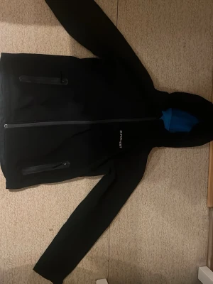 Svart vindjacka från Everest - Snygg svart vindjacka från Everest med blått innerfoder i huvan. Jackan har dragkedja framtill och två praktiska bröstfickor med dragkedja. Perfekt för blåsiga dagar och enkel att matcha med olika outfits.