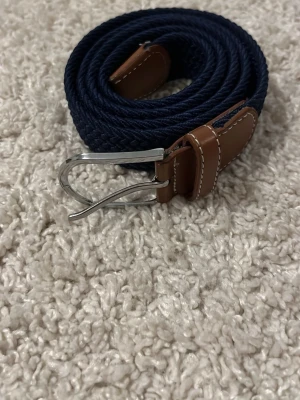 Mörkblått flätat bälte med läderdetaljer - Snyggt mörkblått flätat bälte med bruna läderdetaljer och silverfärgat spänne. Bältet har en stilren design och passar perfekt till jeans eller chinos. Materialet är en mix av textil och läder vilket ger både komfort och stil.