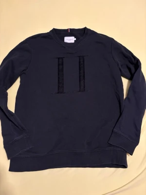 Svart sweatshirt från Les Deux - Snygg svart sweatshirt från Les Deux med broderad II-logga på bröstet. Tröjan har rund halsringning, ribbade muddar vid ärmslut och nederkant samt en diskret detalj med röd och vit rand i nacken. Perfekt för en clean och stilren look.