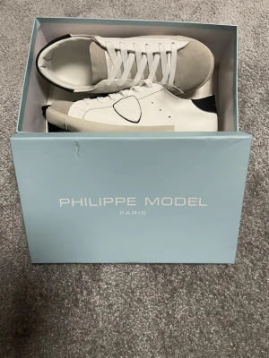 Vita Philippe Model sneakers - Snygga vita sneakers från Philippe Model med grå mockadetaljer på tån och svart hälparti. Klassisk låg modell med snörning och rund tå. Skorna har en stilren design och är tillverkade i skinn och mocka för en exklusiv känsla. Pris kan diskuteras vid snabb affär! 