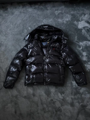Svart pufferjacka från Polo Ralph Lauren - Svart glansig pufferjacka från Polo Ralph Lauren med huva och dragkedja framtill. Jackan har en quiltad design och Polo-logga broderad i rött på bröstet. Perfekt för kalla dagar med sin varma och stilrena look.