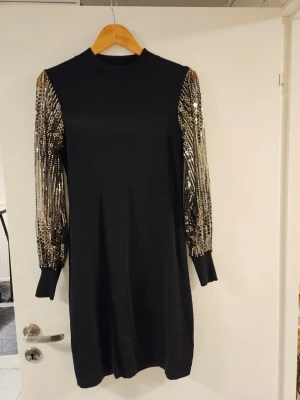 Svart klänning med paljettärmar - Svart klänning med långa ärmar täckta av guldiga paljetter som ger en riktigt snygg kontrast. Rak passform och ribbad halsringning. Perfekt för dig som vill sticka ut med coola detaljer och glittriga ärmar.