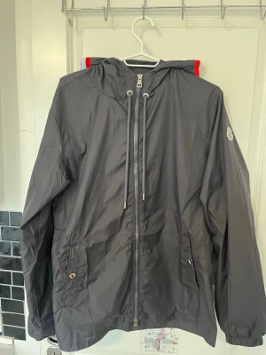 Moncler Windbreaker  - Snygg svart vindjacka från Moncler med huva och dragsko. Jackan har dragkedja framtill, två fickor med knapp och Moncler-logga på ärmen. Perfekt för dig som vill ha en stilren och funktionell jacka med exklusiv känsla. Storlek 3 sitter som M/L