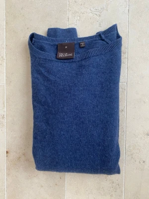 Blå stickad tröja från Oscar Jacobson - Mysig blå stickad tröja från Oscar Jacobson i mjukt material. Tröjan har rund halsringning, långa ärmar och ribbade muddar vid ärmslut och nederkant. Perfekt för lager på lager och enkel att matcha med jeans eller chinos.