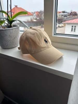 Beige keps från Polo Ralph Lauren - Snygg beige keps från Polo Ralph Lauren med klassisk böjd skärm och justerbar rem bak. Kepsen har den ikoniska broderade loggan med en svart häst på framsidan. Tillverkad i bomull för en skön och avslappnad look.