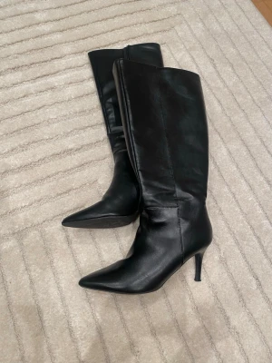 Svarta stövlar från Nelly  med klack och spetsig tå - Snygga svarta stövlar i skinn med spetsig tå och smal hög klack. Klassisk och stilren modell som går upp till knäet. Dragkedja på insidan för enkel påtagning. Perfekta för att lyfta vilken outfit som helst.