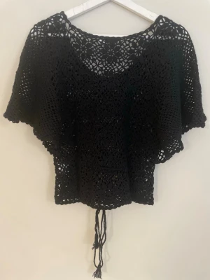 Svart virkad croppad topp från Popin - Svart virkad topp från Poony med croppad passform och knytband nertill. Toppen har korta ärmar och ett luftigt, mönstrat virkat material som ger en bohemisk känsla. Perfekt att styla över ett linne eller bikini.