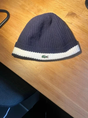 Mörkblå ribbstickad mössa Lacoste - Säljer en mörkblå ribbstickad mössa från Lacoste med vit kant och den klassiska gröna krokodilloggan framtill. Mössan har en enkel, stilren design och är perfekt för dig som vill ha en snygg accessoar till hösten och vintern.