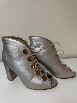 Silvriga stövletter med snörning och klack - Unika silvriga stövletter med snörning framtill och stora runda guldfärgade knappar. Skorna har öppen tå och hög klack, vilket ger en cool retrovibe. Tillverkade i skinnliknande material och perfekta för dig som vill sticka ut.