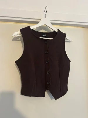Mörkbrun väst med knappar - Snygg mörkbrun väst från Gina Tricot med fem knappar framtill. Västen har en figurnära passform och är ärmlös med rundad halsringning. Perfekt att styla över en skjorta eller t-shirt för en trendig look.