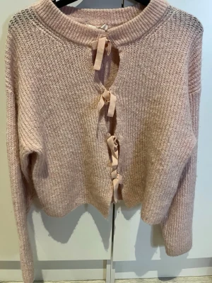 Rosa stickad kofta med rosetter Gina Tricot - Supersöt ljusrosa stickad kofta från Gina Tricot med fyra dekorativa rosetter framtill istället för knappar. Koftan har lång ärm, bred ribbad krage och är gjord i sopft, mysigt material. Perfekt för lager på lager och en gullig touch till din outfit.