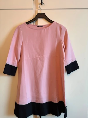 Rosa och svart klänning - Helt ny, rosa klänning med svarta detaljer vid ärmslut och nederkant. Klänningen har rund halsringning och trekvartsärm. Storlek XS, men passar också en S
