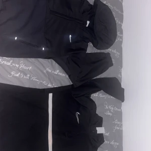 Svart Nike vindjacka med huva - Snygg svart vindjacka från Nike med huva och dragkedja framtill. Jackan har reflekterande detaljer och en minimalistisk logga på bröstet. Perfekt för blåsiga dagar och enkel att matcha med olika outfits.