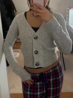 Ljusgrå cropped kofta med stora knappar - Superfin ljusgrå cropped kofta med stora, mörka knappar framtill. Koftan har v-ringning, långa ärmar och är stickad i ett mjukt material. Perfekt att slänga över ett linne eller t-shirt för en chill look.