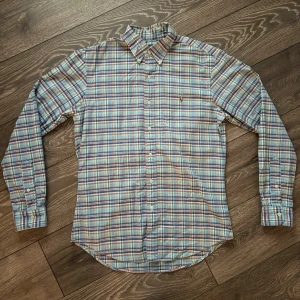 Rutig skjorta från Ralph lauren i bomull - Klassisk rutig skjorta från Ralph lauren i mjuk bomull. Skjortan har knappar framtill, button-down krage och långa ärmar med manschett. Färgerna går i blått, grönt, vitt och rött. Perfekt för en avslappnad men ändå stilsäker look.