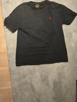 Svart t-shirt från Polo Ralph Lauren - Klassisk svart t-shirt från Polo Ralph Lauren med korta ärmar och rund hals. T-shirten har den ikoniska röda logotypen broderad på bröstet. Tillverkad i mjuk bomull för en bekväm känsla och enkel stil.