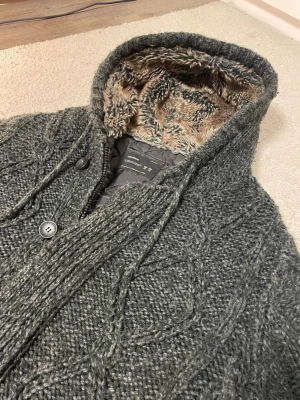 Mörkgrå stickad hoodie med fuskpäls - Mysig svartgrå stickad hoodie med knäppning och dragkedja framtill och fuskpälsfodrad huva. Tröjan har kabelstickat mönster, ribbade muddar och två fickor framtill. Perfekt för kalla dagar och riktigt skön att bära🧊 Den fungerar både som jacka och hoodie! Hör av er vid intresse! Pris är ej hugget i sten🪨 