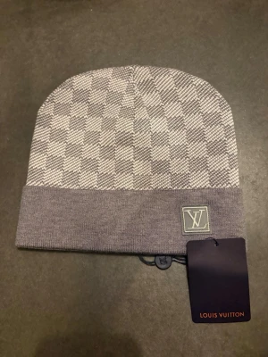 Grå exklusiv mössa med rutmönster - Snygg grå mössa från Lui ton med klassiskt rutmönster och vikt kant. Lui ton- framtill och diskret etikett på insidan. Perfekt accessoar för dig som gillar exklusiv streetstyle och vill sticka ut med lyxig design.