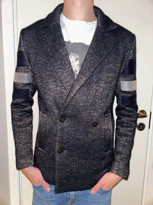 Mörkblå skepparkavaj med ränder - Snygg mörkblå skepparkavaj med breda grå och blå ränder på ärmar och rygg. Dubbelknäppt framtill med fyra knappar och klassisk krage. Tillverkad i mjuk ullblandning som ger en skön känsla och stilren look. Perfekt för dig som vill sticka ut lite extra.