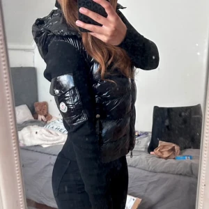 Svart glansig dunväst från Moncler XS/S  - Säljer en svart glansig dunväst kardigan  från Moncler med huva och ikonisk logga på fickan. Kardigan har en figurnära passform och praktiska fickor med lock. Perfekt för lager-på-lager och ger en cool streetstyle-look.