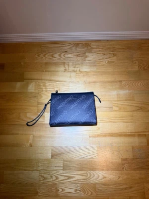 Svart kuvertväska från Valentino - Stilren svart kuvertväska från Valentino med logotypmönster över hela väskan. Väskan har en avtagbar handledsrem och dragkedja upptill. Perfekt för att bära det viktigaste och ger en lyxig känsla till din outfit.