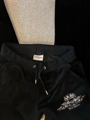 Svarta velourbyxor från Juicy Couture - Mysiga svarta velourbyxor från Juicy Couture med dragsko i midjan och silverfärgade detaljer. Byxorna har ett broderat emblem på ena benet och är perfekta för chill dagar hemma eller när du vill vara extra comfy.