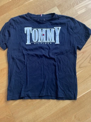 Svart Tommy Hilfiger t-shirt - Snygg svart t-shirt från Tommy Hilfiger med stort broderat TOMMY HILFIGER-logo i vitt på bröstet. Klassisk rund hals och korta ärmar. Perfekt för dig som gillar streetwear och vill ha en enkel men ikonisk look.