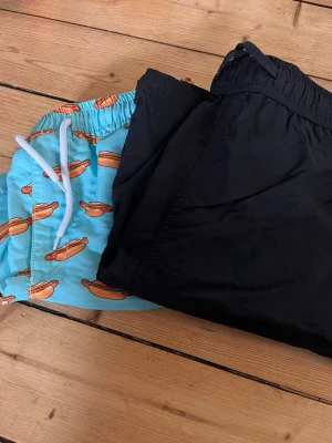 Två par badbyxor, svart & hotdog-mönster - Säljer två par badbyxor, ett par helsvarta och ett par ljusblå med biltema mönster. Båda har elastisk midja och klassisk passform. Materialet känns lätt och snabbtorkande. Knappt användanochbi jätte fint skick. 100kr för båda