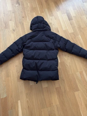 Svart pufferjacka från Sailracing - Svart pufferjacka från sail racing. Storlek 150 med huva och dragkedja framtill. Jackan har två sidofickor med dragkedja och logga på bröstet. Tjockt vadderad för extra värme och har en klassisk, rak passform. Perfekt för kalla dagar. Jag är 167 och den är lite för liten. Alltså ganska stor i storleken 